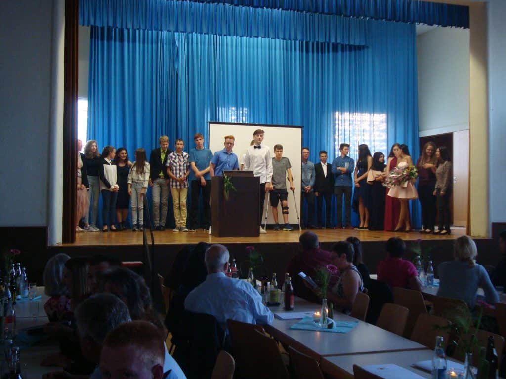 Auf einer Bühne mit blauem Vorhang steht eine Gruppe von Menschen, einige in festlicher Kleidung, um ein Podium versammelt. Im Vordergrund sitzen Menschen an Tischen mit Getränken und Kerzen und beobachten die Bühne.