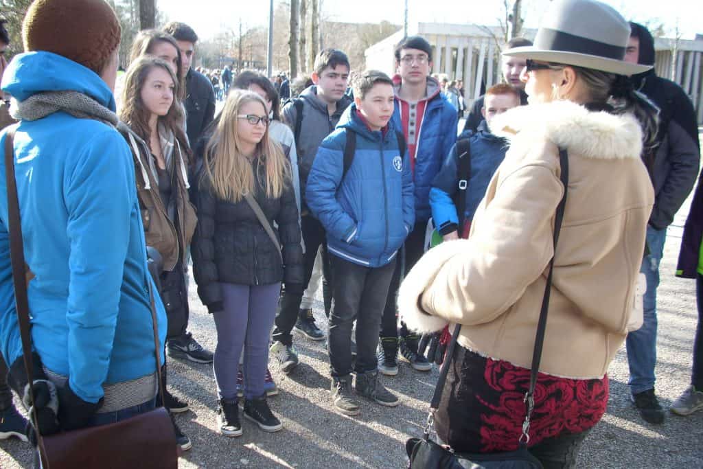 Eine Gruppe Jugendlicher in Winterjacken steht im Halbkreis im Freien und hört einem Erwachsenen zu, der einen hellbraunen Mantel mit Pelzkragen und eine graue Mütze trägt. Im Hintergrund sind Bäume und ein Gebäude zu sehen.