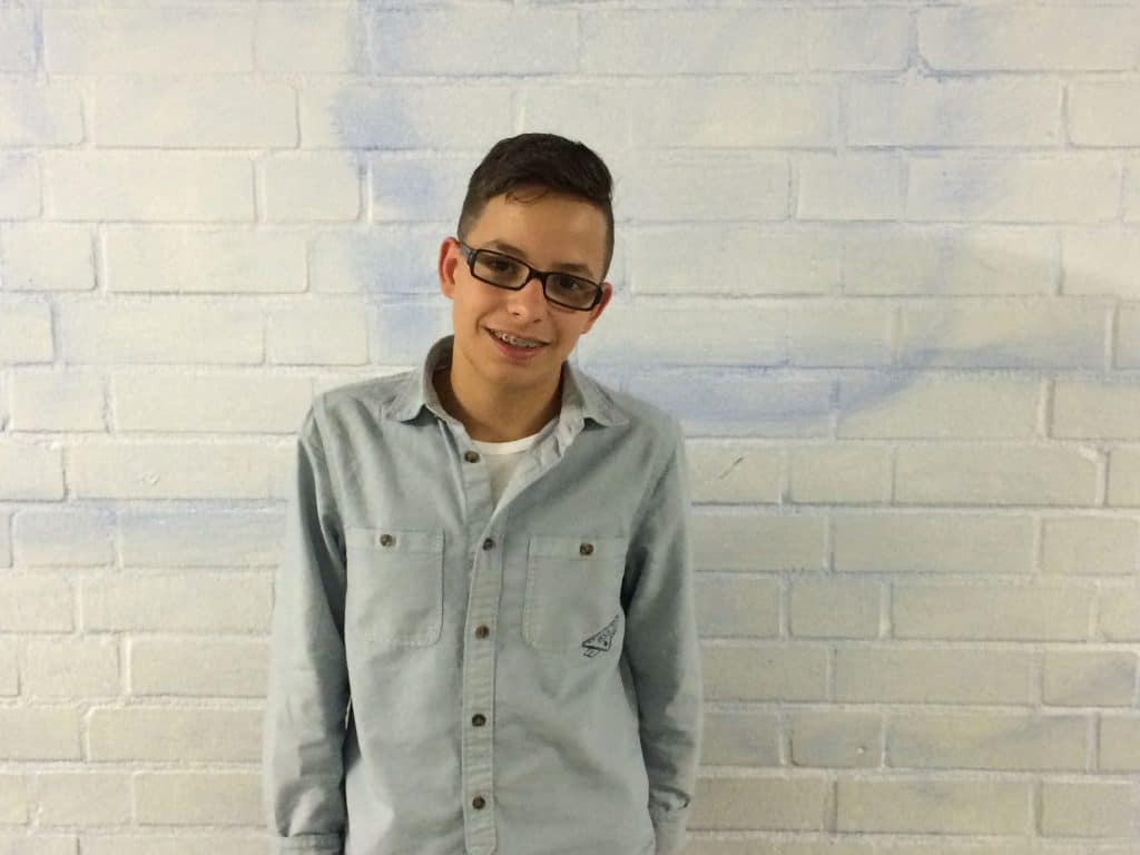Ein Teenager mit kurzen dunklen Haaren, Brille und Zahnspange steht lächelnd vor einer weißen Backsteinwand. Er trägt ein hellblaues Hemd über einem weißen T-Shirt.