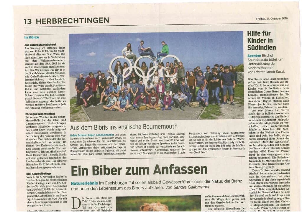 Eine große Gruppe von Menschen posiert vor der Skulptur der Olympischen Ringe im Freien auf einem grasbewachsenen Hügel, mit leicht bewölktem Himmel und einer fernen Landschaft im Hintergrund. Das Foto erschien in einer deutschen Zeitung.