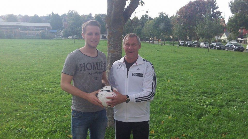 Zwei Männer stehen auf einer Wiese. Einer trägt ein graues T-Shirt und Jeans, der andere ein weißes Sakko. Sie lächeln und halten gemeinsam einen Fußball in den Händen. Im Hintergrund sind Bäume und parkende Autos zu sehen.