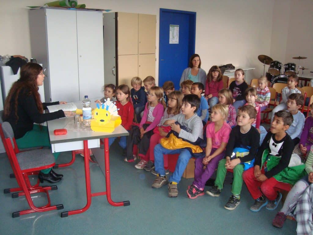 Eine Frau sitzt an einem Schreibtisch und liest einer Gruppe kleiner Kinder vor, die in Reihen in einem Klassenzimmer sitzen und aufmerksam zuhören. Im Hintergrund sind Lehrer, Schränke und Musikinstrumente zu hören.