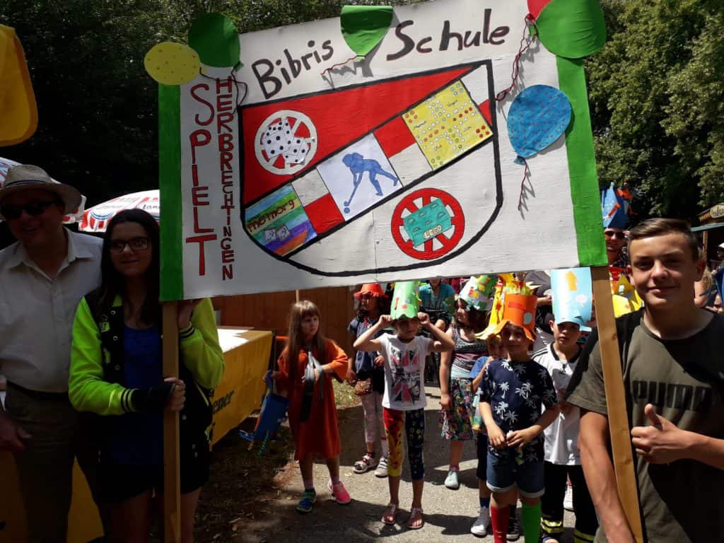 Eine Gruppe von Kindern und Erwachsenen steht draußen und hält ein buntes, handgemachtes Schild mit der Aufschrift „Bibis Schule“ sowie Zeichnungen und Dekorationen hoch. Einige Kinder tragen Hüte und Kostüme. Im Hintergrund sind Bäume zu sehen.
