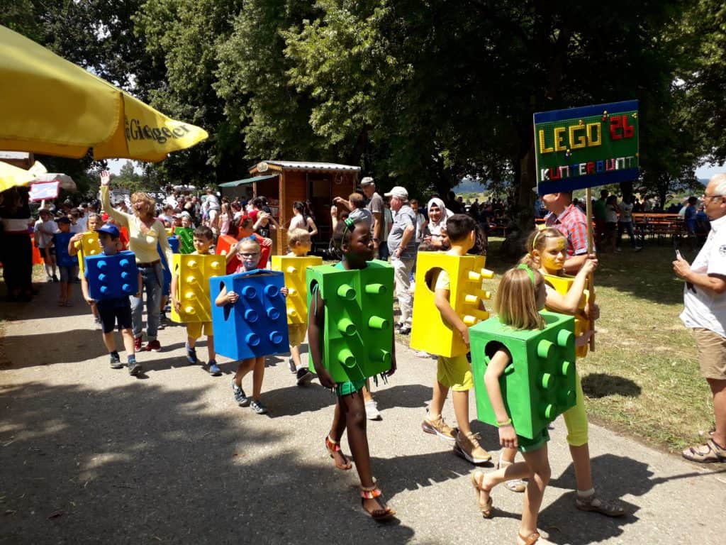 Eine Gruppe von Kindern, die als riesige LEGO-Steine in den Farben Blau, Grün und Gelb verkleidet sind, zieht bei einem Fest oder einer Veranstaltung im Freien umher und hält ein buntes Schild mit der Aufschrift „LEGO®-AG Kinderumzug“, umgeben von Menschen und Bäumen.