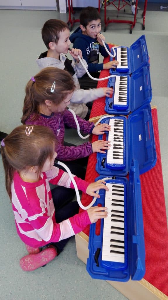 Vier kleine Kinder sitzen in einer Reihe und spielen Melodicas. Jedes Kind hat ein blaues Gehäuse und weiße Tasten. Drei Mädchen und ein Junge benutzen Mundstücke, die über Schläuche mit den Instrumenten verbunden sind. Sie konzentrieren sich auf das gemeinsame Musizieren.