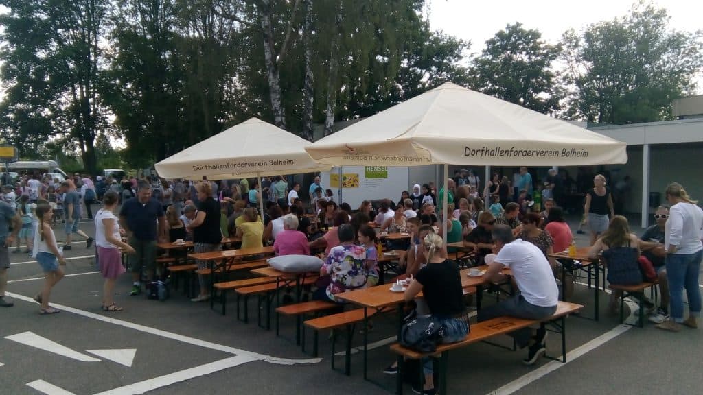 Eine große Gruppe von Menschen hat sich im Freien versammelt. Sie sitzen an Holztischen unter zwei weißen Sonnenschirmen und genießen Essen und Getränke bei einer Gemeinschaftsveranstaltung. Im Hintergrund sind Bäume und andere Besucher zu sehen.