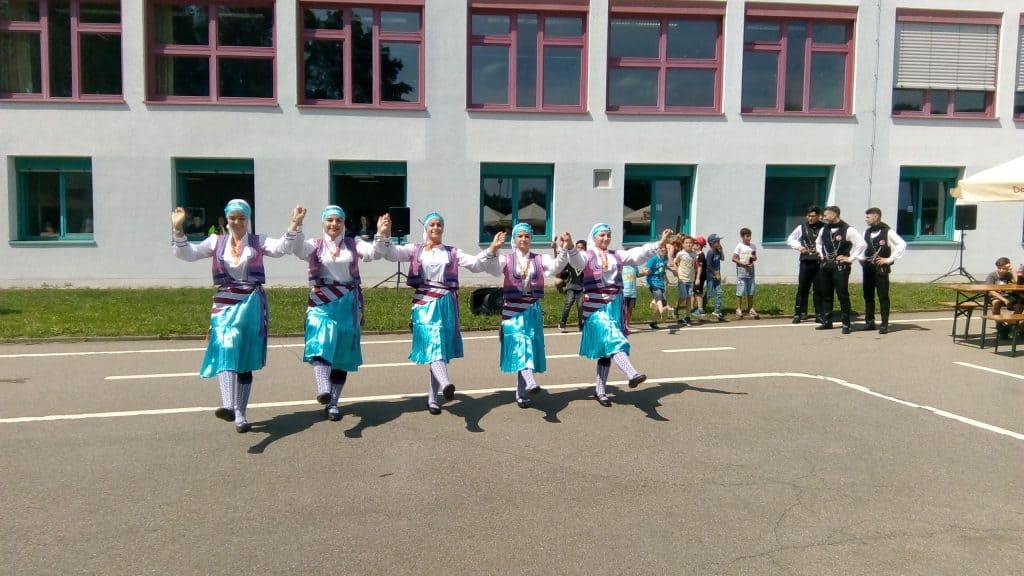 Fünf Personen in passenden traditionellen blauen und violetten Outfits führen im Freien vor einem Gebäude einen Gruppentanz auf, während im Hintergrund ein kleines Publikum zusieht.
