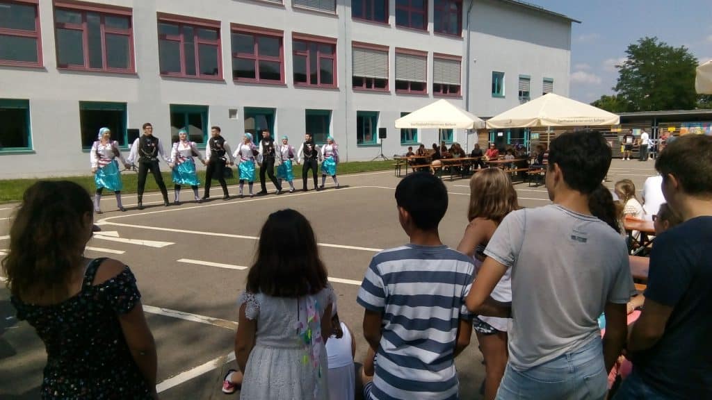 Eine Gruppe von Menschen führt vor einem Gebäude einen Tanz auf, während ein Publikum aus Kindern und Erwachsenen im Vordergrund und von Tischen unter Sonnenschirmen draußen zusieht.