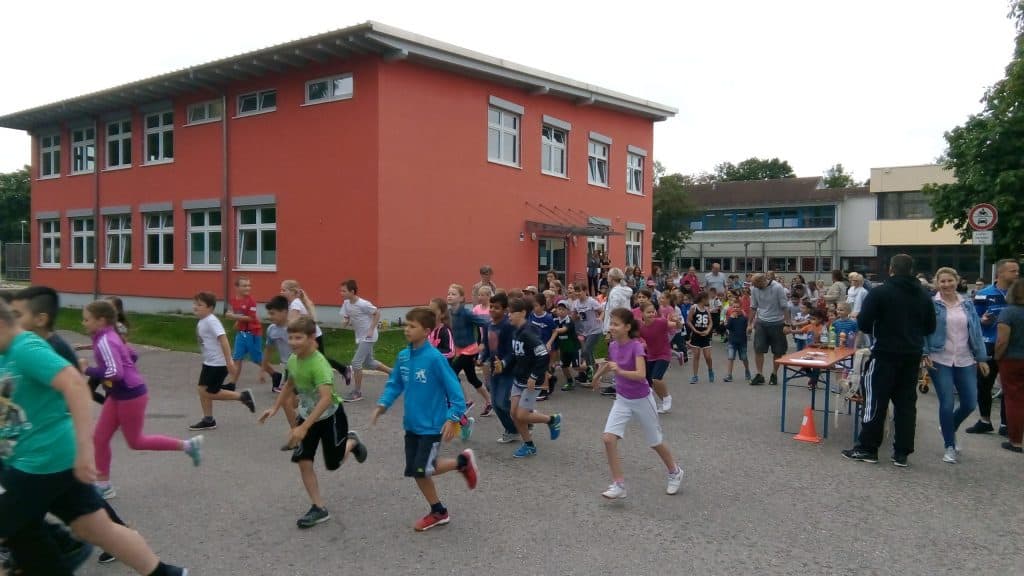Eine Gruppe Kinder nimmt an einem Rennen in der Nähe eines roten Gebäudes teil und wird dabei von Erwachsenen und Passanten beobachtet. Einige Kinder rennen, während andere auf den Start warten. So entsteht eine lebhafte Szene im Freien.