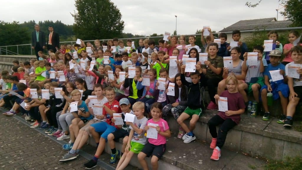 Eine große Gruppe Kinder sitzt auf einer Außentreppe, hält Zertifikate hoch und lächelt in die Kamera. Einige Erwachsene stehen dahinter. Im Hintergrund sind Bäume und Gebäude unter einem bewölkten Himmel zu sehen.