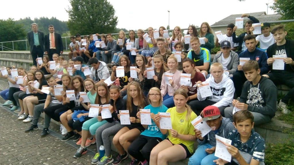 Eine große Gruppe von Studierenden sitzt draußen auf einer Treppe, hält Zertifikate in den Händen und lächelt in die Kamera. Zwei Erwachsene stehen im Hintergrund. Im Hintergrund sind Bäume und Gebäude zu sehen.