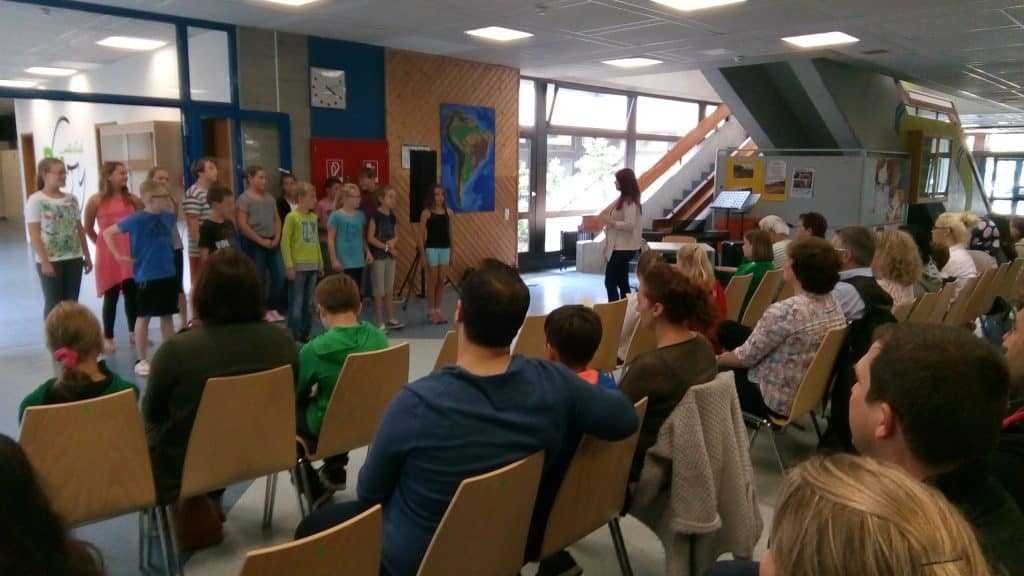 Eine Gruppe von Kindern steht in einer Reihe und führt vor einem Publikum aus sitzenden Erwachsenen und Kindern in einem hell erleuchteten Raum mit großen Fenstern und bunten Wänden eine Aufführung oder Präsentation auf.