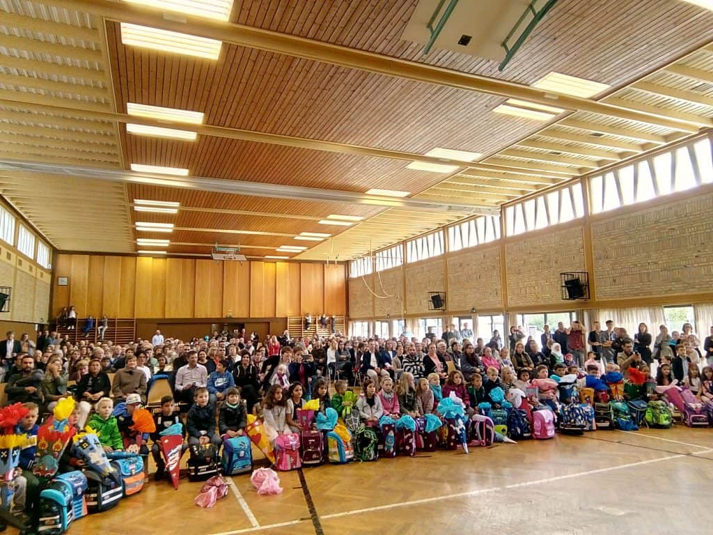 Eine große Gruppe Kinder mit bunten Rucksäcken sitzt in der ersten Reihe einer überfüllten Turnhalle. Viele Erwachsene sitzen dahinter, alle mit dem Blick nach vorne gerichtet, in einem gut beleuchteten Raum mit Wänden und Decke aus Holz.