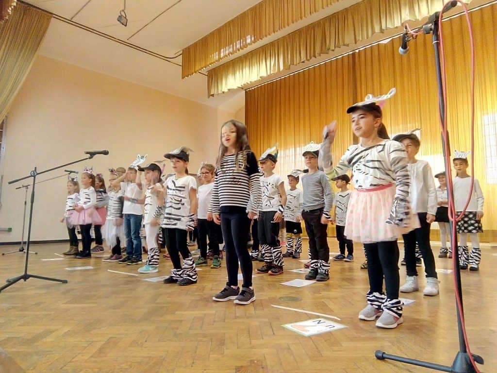 Eine Gruppe von Kindern in Tierkostümen, überwiegend weiß mit schwarzen Streifen, steht in Reihen auf einer Holzbühne in einem hell erleuchteten Raum mit gelben Vorhängen und tritt auf oder singt in Mikrofone.