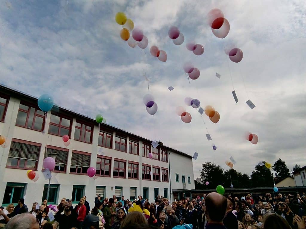 Eine große Gruppe von Menschen versammelt sich vor einem Schulgebäude und beobachtet, wie bunte Ballons mit daran befestigten Notizen in den bewölkten Himmel aufsteigen.