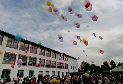 Eine große Gruppe von Menschen versammelt sich vor einem Schulgebäude und beobachtet, wie bunte Ballons mit daran befestigten Notizen in den bewölkten Himmel aufsteigen.