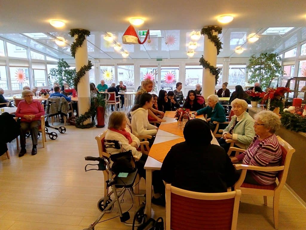 Eine Gruppe älterer Menschen und Pflegekräfte sitzt in einem hellen, festlichen Gemeinschaftsraum an einem dekorierten Tisch, knüpft Kontakte und genießt die Gesellschaft der anderen. Weihnachtsdekorationen und Grünpflanzen schmücken den Raum.