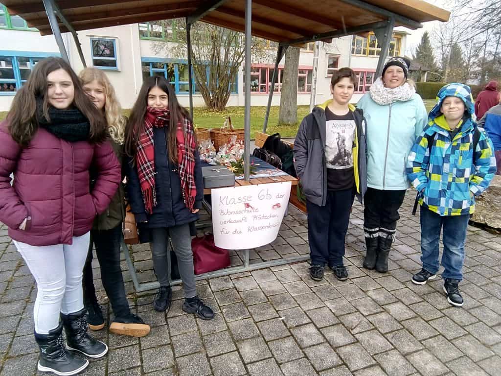 Sechs Kinder stehen lächelnd vor einem Tisch, an dem Kunsthandwerk verkauft wird. Ein Schild auf dem Tisch weist auf Deutsch darauf hin, dass die Klasse 6b Kunsthandwerk für einen wohltätigen Zweck verkauft. Die Szene spielt auf einem Schulhof im Freien.