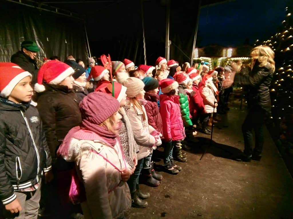 Ein Kinderchor in Winterkleidung und Weihnachtsmannmützen singt nachts unter der Leitung eines Dirigenten auf einer Bühne. Im Hintergrund sind festliche Lichter und Dekorationen zu sehen.