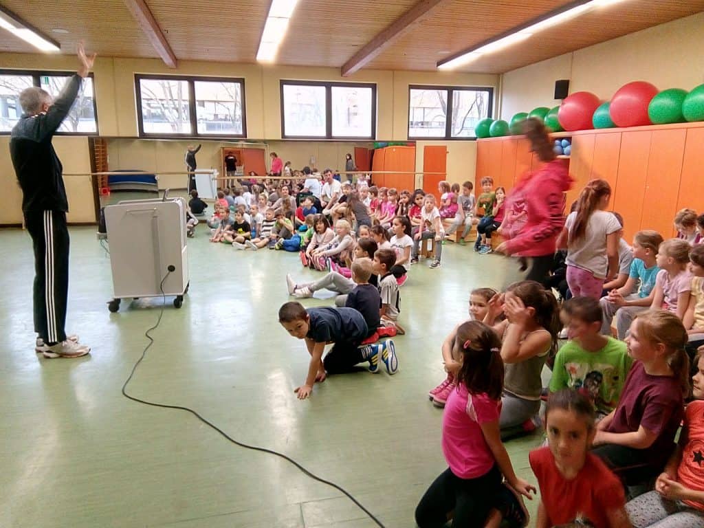 Eine große Gruppe Kinder sitzt in einer Turnhalle auf dem Boden, einige Erwachsene stehen daneben. Ein Erwachsener vorne hebt die Hand, während bunte Gymnastikbälle in Regalen an der Wand aufgereiht sind. Die Szene wirkt lebendig und energiegeladen.