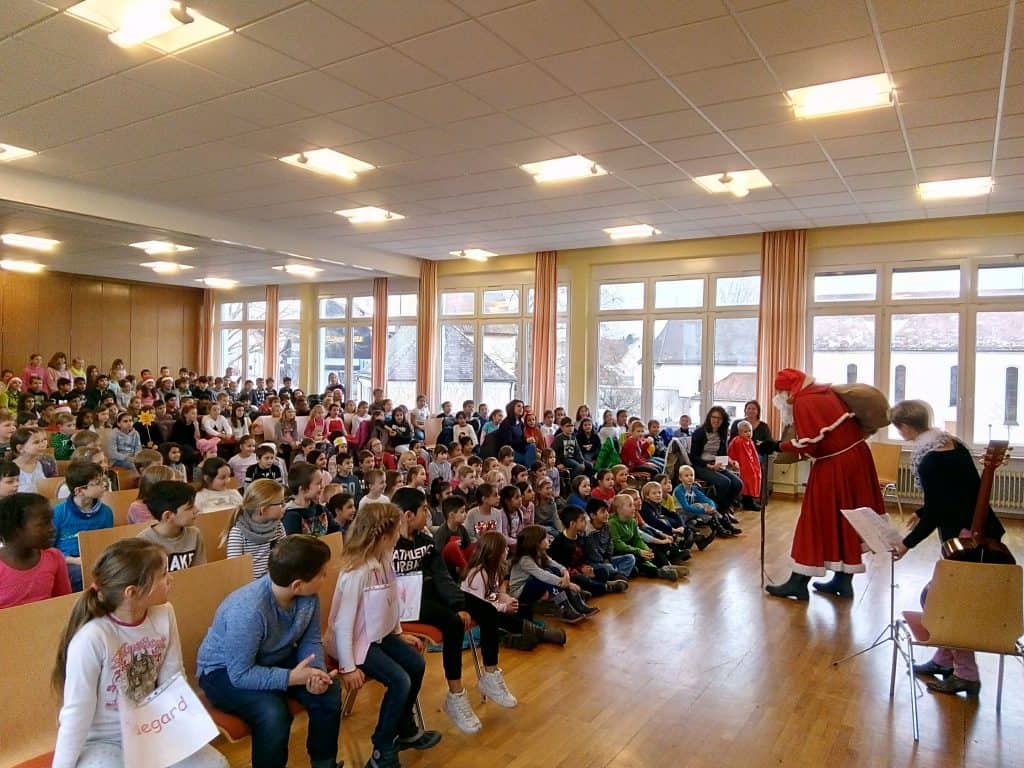 Eine große Gruppe Kinder sitzt in einer Schulaula und beobachtet eine als Nikolaus verkleidete Person und eine weitere Person im Kostüm in der ersten Reihe. Erwachsene stehen daneben, und durch die hohen Fenster fällt natürliches Licht in den Raum.