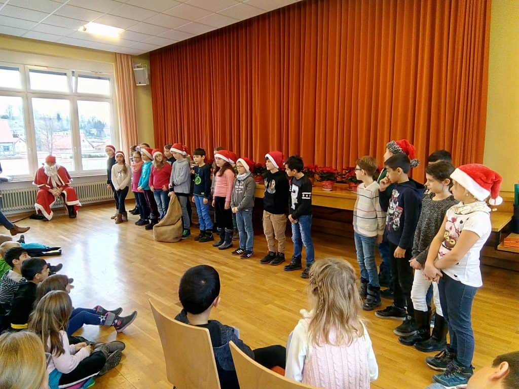 Eine Gruppe von Kindern mit Weihnachtsmannmützen steht in einer Reihe auf der Bühne und tritt vor einem sitzenden Kinderpublikum auf. Zwei als Weihnachtsmann verkleidete Erwachsene sitzen am Fenster. Der Raum hat Holzböden und orangefarbene Vorhänge.