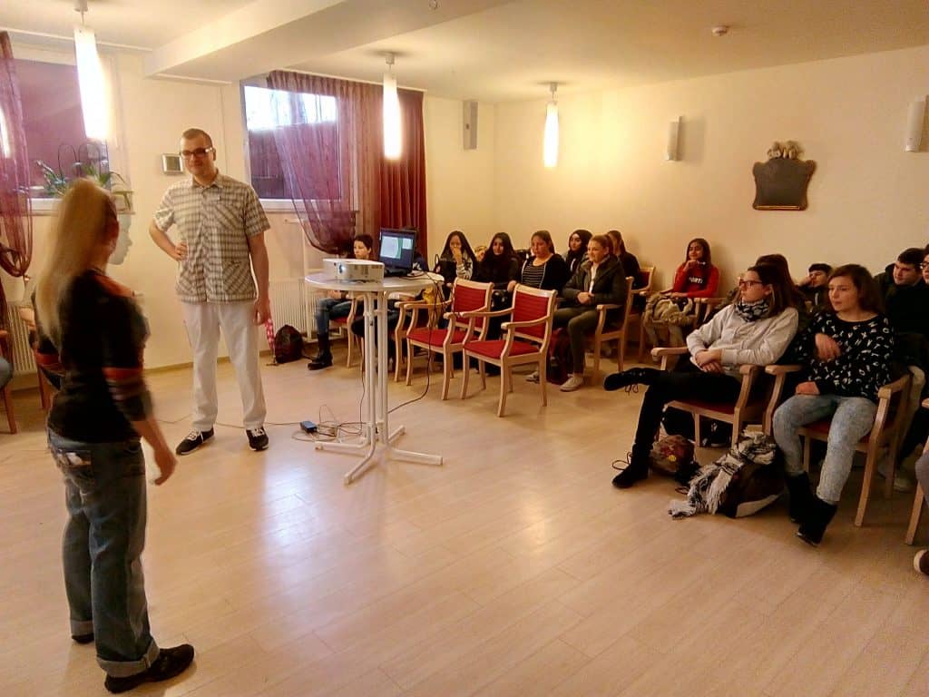 Eine Frau steht einem Mann gegenüber, der neben einem Projektor steht. Eine Gruppe von Menschen sitzt auf Stühlen und beobachtet sie in einem hell erleuchteten Raum. Auf einem kleinen Tisch steht ein Laptop, im Hintergrund hängen kastanienbraune Vorhänge.