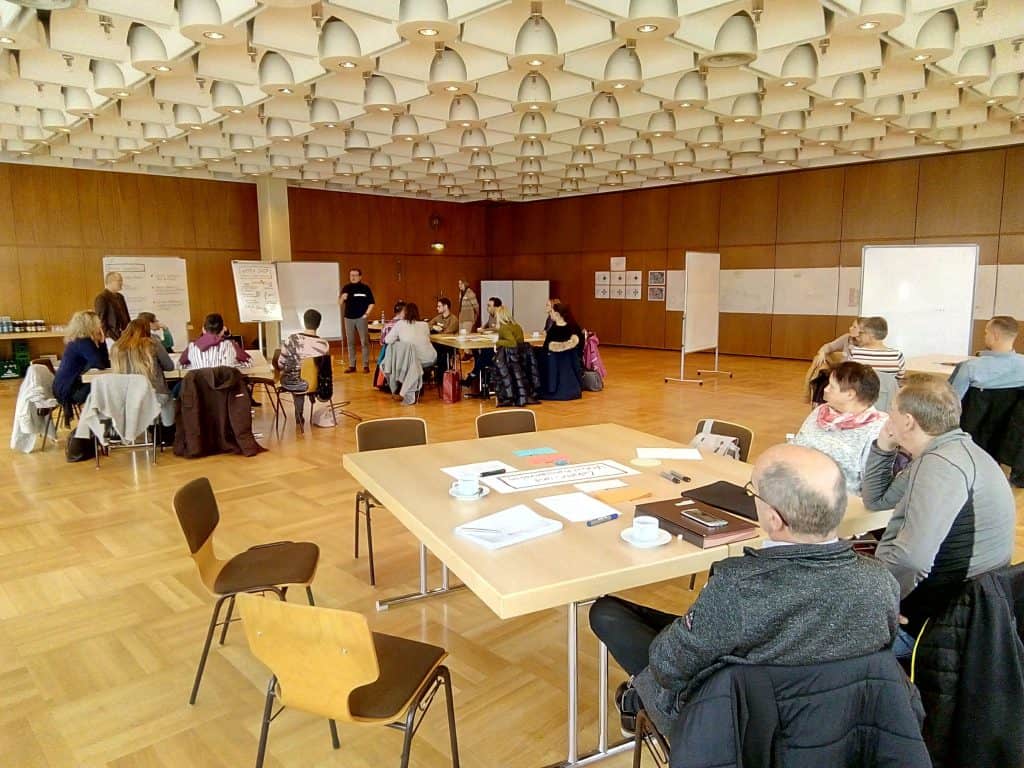 Eine Gruppe von Menschen sitzt in einem großen, modernen Konferenzraum mit Holzboden und geometrischen Deckenpaneelen an Tischen für einen Workshop oder ein Seminar. Überall im Raum sind Whiteboards und Flipcharts aufgestellt.