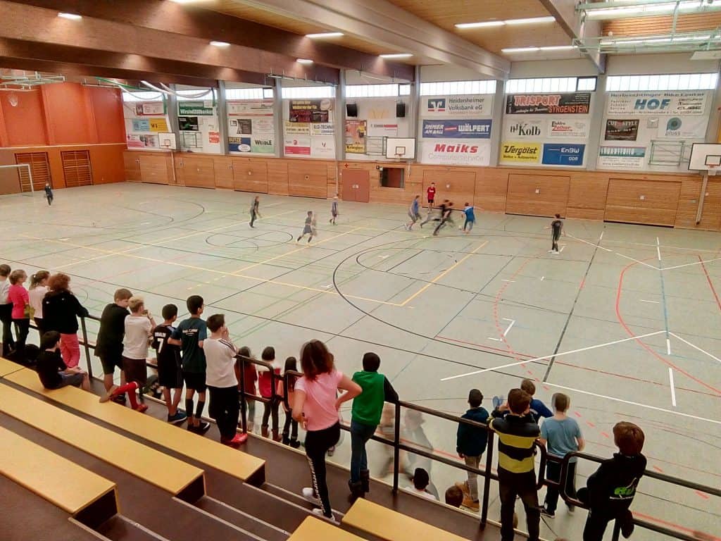 Kinder spielen in einer großen Turnhalle Hallenfußball, während andere von der Tribüne aus zuschauen. Die Turnhalle hat hohe Decken, Holzwände und über dem Spielfeld hängen Werbebanner.