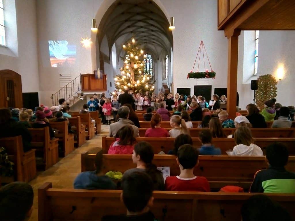 Eine Gruppe von Kindern singt vor einem geschmückten Weihnachtsbaum in einer Kirche, während das Publikum in Holzbänken sitzt und der Aufführung zusieht. Die Kirche ist hell mit gewölbten Decken und festlicher Dekoration.