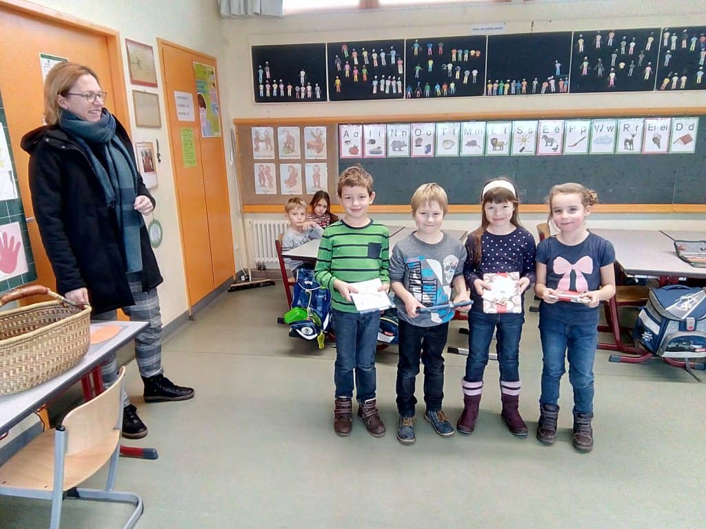 Fünf kleine Kinder stehen in einem Klassenzimmer und halten Geschenke in den Händen. Sie lächeln in die Kamera. Links steht eine Frau und beobachtet sie. Andere Kinder sitzen im Hintergrund. An den Wänden des Klassenzimmers hängen Zeichnungen und Kunstwerke.