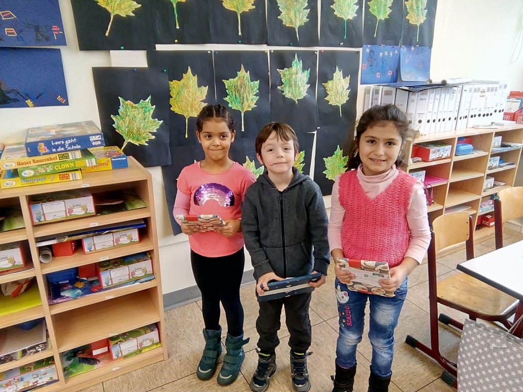 Drei kleine Kinder stehen lächelnd in einem Klassenzimmer und halten jeweils ein Buch in der Hand. Hinter ihnen hängt ein Blattbild an der Wand, und links sind Regale mit Spielen und Büchern zu sehen.