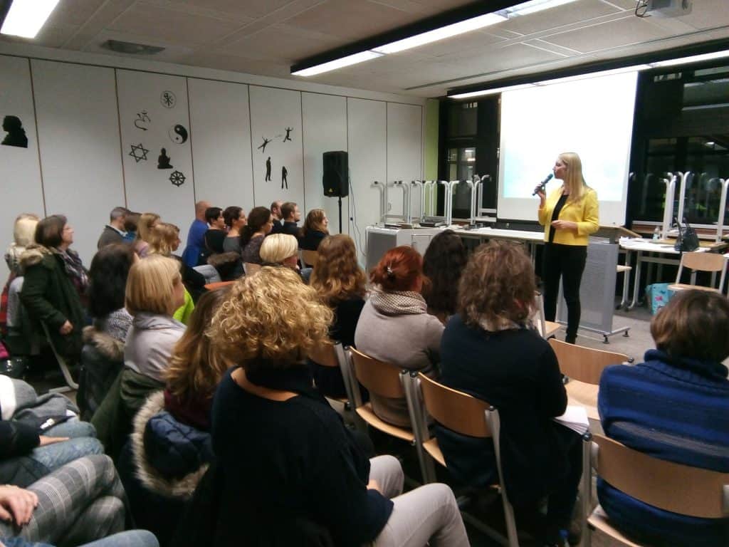 Eine Frau in einer gelben Jacke spricht vor einem Publikum in einem Klassenzimmer oder Hörsaal in ein Mikrofon. Im Hintergrund sind eine Projektionsleinwand und gestapelte Stühle zu sehen.