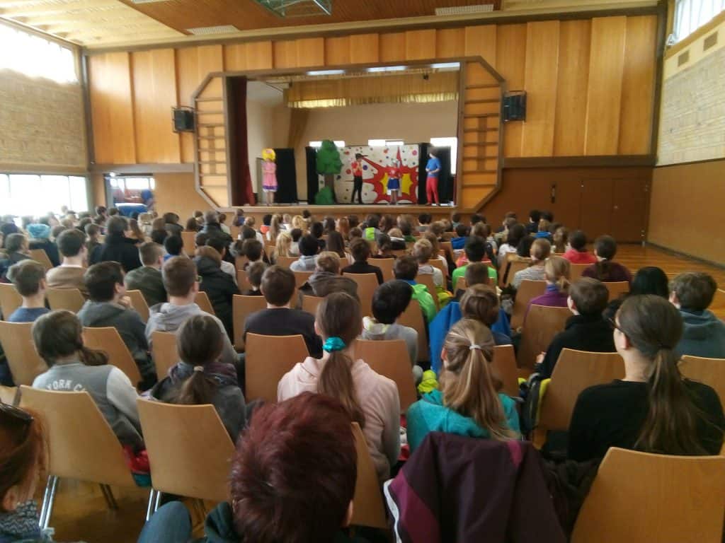 Eine große Gruppe Kinder sitzt in Stuhlreihen und sieht sich eine farbenfrohe Bühnenaufführung in einer Schulaula mit Holzwänden und hohen Fenstern an. Vier kostümierte Darsteller stehen vor einer dekorierten Kulisse auf der Bühne.