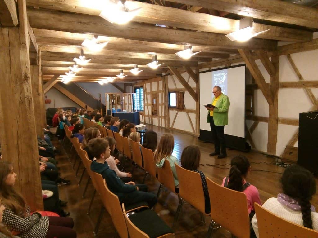 Ein Mann steht in einem rustikalen Raum mit Holzbalken und einer Projektionsleinwand und liest einer Gruppe von Kindern, die in Stuhlreihen sitzen, aus einem Buch vor.