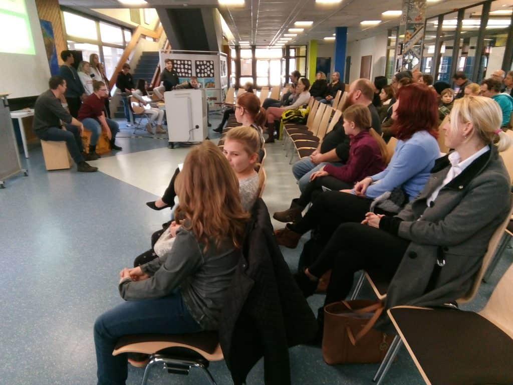 Eine Gruppe von Erwachsenen und Kindern sitzt auf Stühlen in einem hellen, modernen Raum und verfolgt aufmerksam eine Präsentation vorne, wo mehrere Personen vor einer Leinwand stehen.