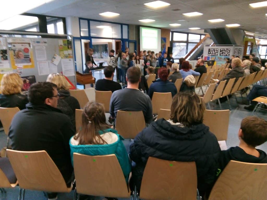 Eine Gruppe von Menschen sitzt auf Stühlen vor einer Bühne, auf der Kinder vor einer Leinwand stehen und möglicherweise etwas aufführen oder präsentieren. Der Raum ist hell und verfügt über Anschlagtafeln und große Fenster.