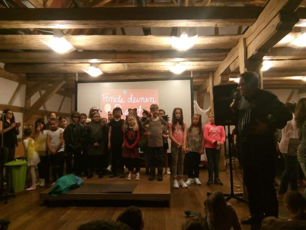 Eine Gruppe kostümierter Kinder steht auf einer Bühne unter Holzbalken und blickt dem Publikum zu. Rechts steht ein Erwachsener mit einem Mikrofon. Auf einem Bildschirm hinter ihnen ist der deutsche Text „Finde deinen Schatz“ zu sehen.