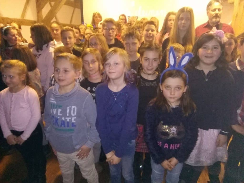 Eine Gruppe von Kindern, einige tragen Stirnbänder mit Hasenohren, steht dicht beieinander in einem Raum und posiert für ein Foto. Hinter den Kindern stehen Erwachsene. Im Hintergrund sind ein Holzbalken und eine Projektionsleinwand zu sehen.