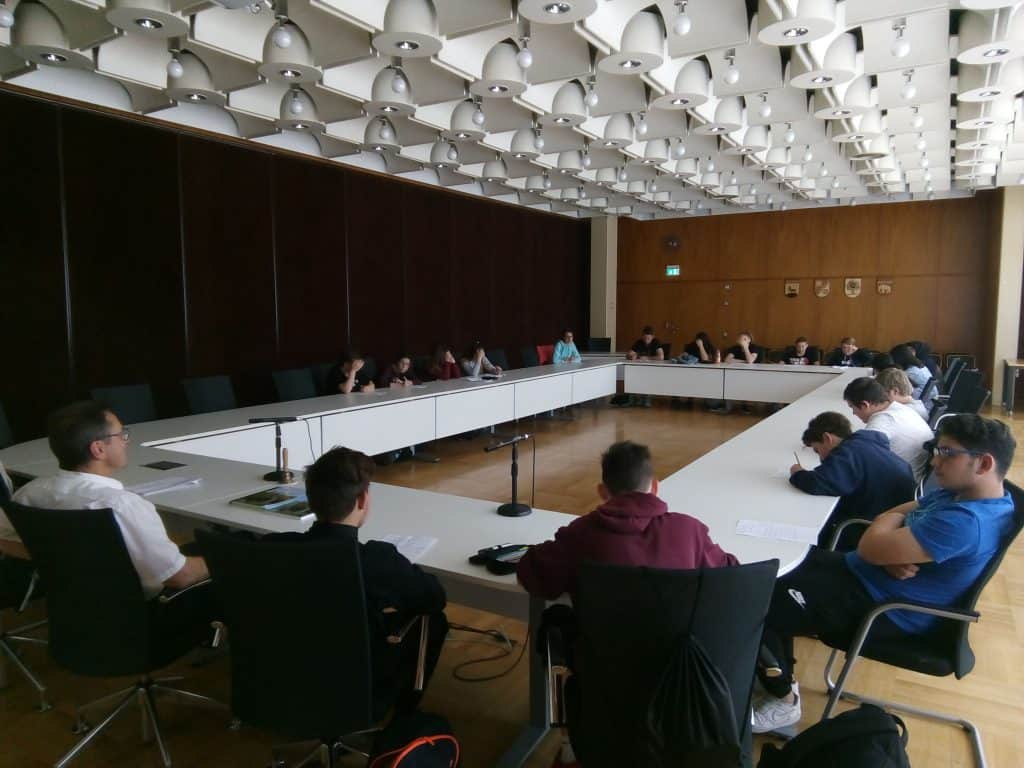 Eine Gruppe von Menschen sitzt um einen großen, rechteckigen Konferenztisch in einem modernen Konferenzraum mit gemusterter Decke und Mikrofonen auf dem Tisch. Einige lesen Dokumente, andere hören aufmerksam zu.