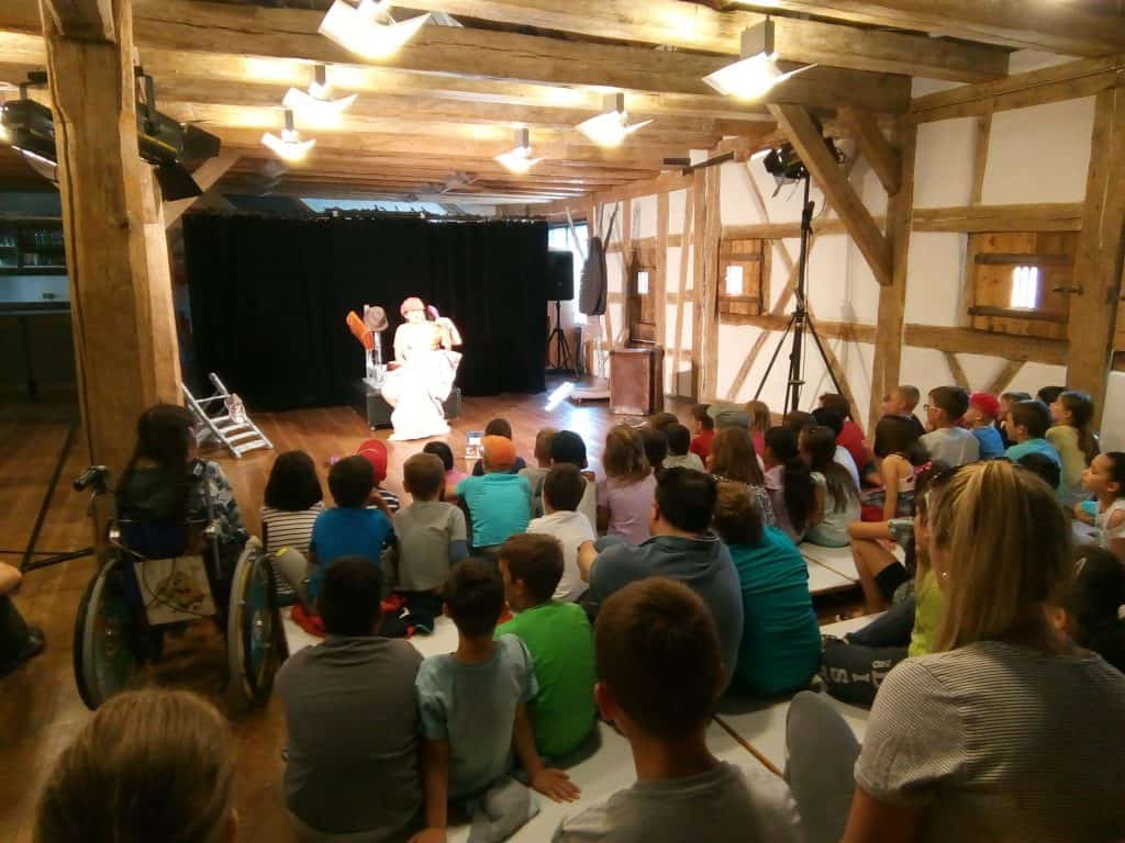 Eine Gruppe Kinder sitzt auf dem Boden und schaut sich eine Bühnenaufführung in einem rustikalen Holzsaal an. Zwei kostümierte Schauspieler treten unter hellem Licht auf. Einige Kinder sitzen auf Matten, eines sitzt im Rollstuhl.