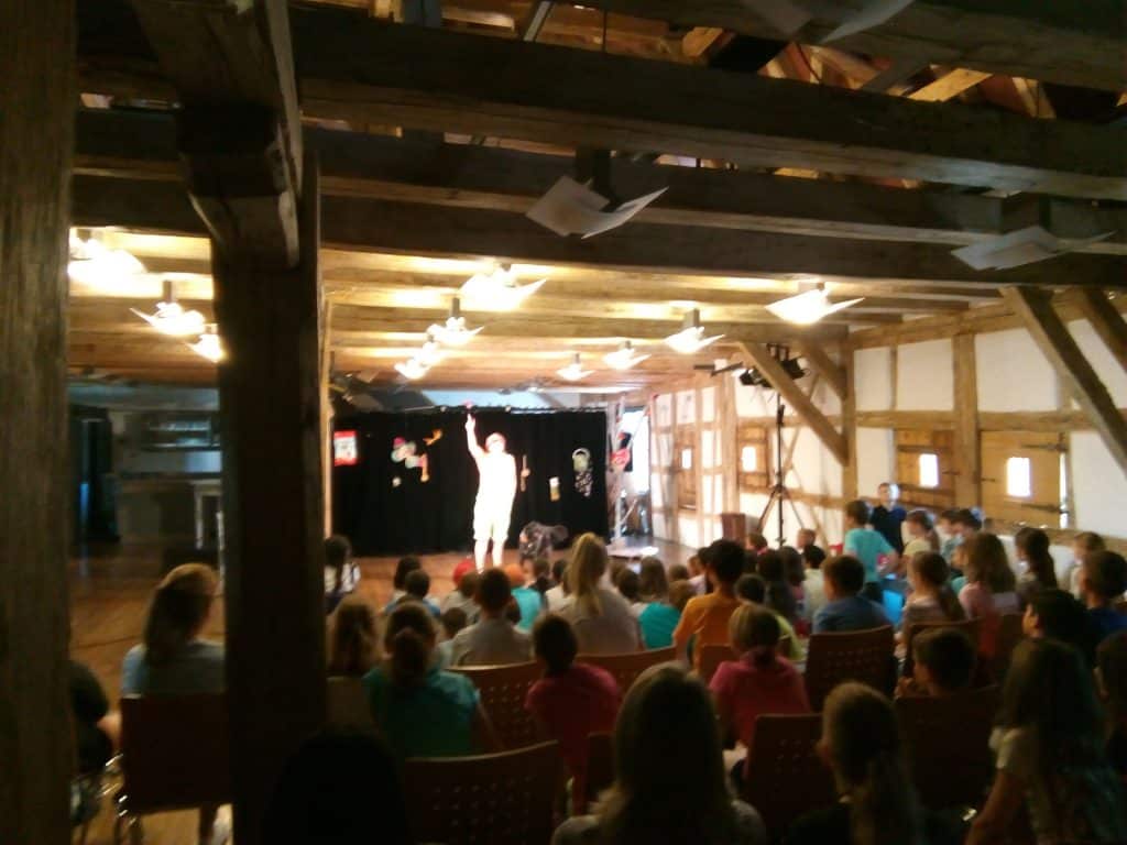 Eine Gruppe Kinder sitzt in Stuhlreihen in einer Holzhalle und beobachtet einen Künstler auf der Bühne unter hellem Licht. Der Künstler steht mit erhobenem Arm da, und das Publikum ist auf die Darbietung konzentriert.