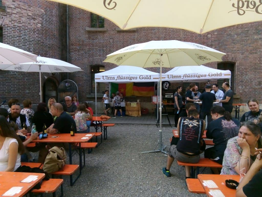 Menschen sitzen und stehen in einem Innenhof an orangefarbenen Picknicktischen unter großen Sonnenschirmen. Einige essen und trinken. Im Hintergrund sind Backsteinmauern und ein Zelt mit einem deutschen Schild zu sehen.