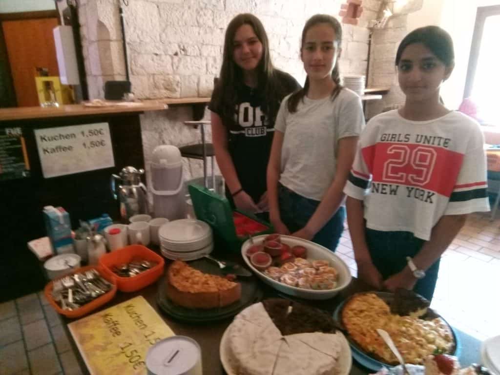 Drei Mädchen stehen in einem gemütlichen Raum hinter einem Tisch mit Kuchen, Gebäck und Kaffee. Auf den Tischen sind die Preise für Kuchen und Kaffee angegeben. Tassen, Teller und Besteck sind ebenfalls ausgestellt.