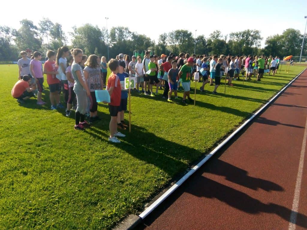 Eine große Gruppe von Menschen, hauptsächlich Kinder und Jugendliche, steht in Reihen auf einer Rasenfläche neben einer Laufbahn und bereitet sich wahrscheinlich auf eine Sportveranstaltung im Freien oder einen sportlichen Wettkampf an einem sonnigen Tag vor.