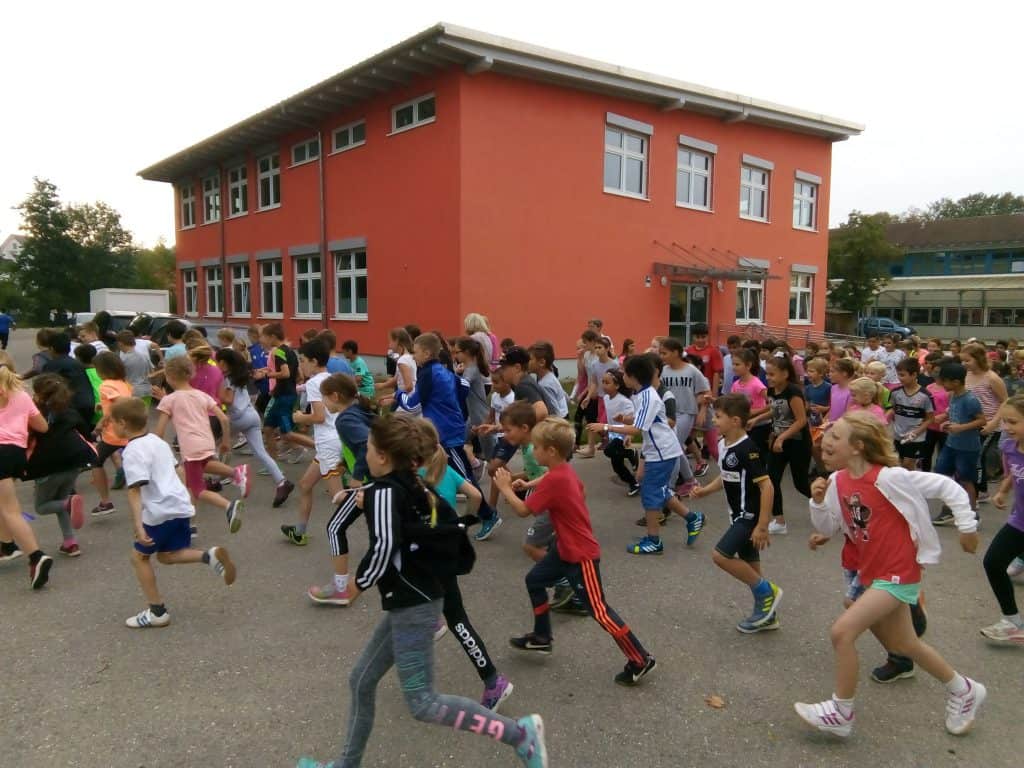 Eine große Gruppe von Kindern läuft gemeinsam im Freien vor einem orangefarbenen Schulgebäude und nimmt anscheinend an einem Schulrennen oder einer Schulveranstaltung teil.