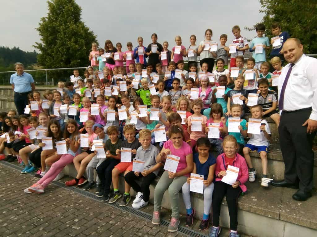 Eine große Gruppe von Kindern sitzt und steht auf einer Außentreppe, hält Zertifikate in den Händen und lächelt in die Kamera. Zwei Erwachsene stehen zu beiden Seiten der Gruppe. Im Hintergrund sind Bäume und Grünflächen zu sehen.