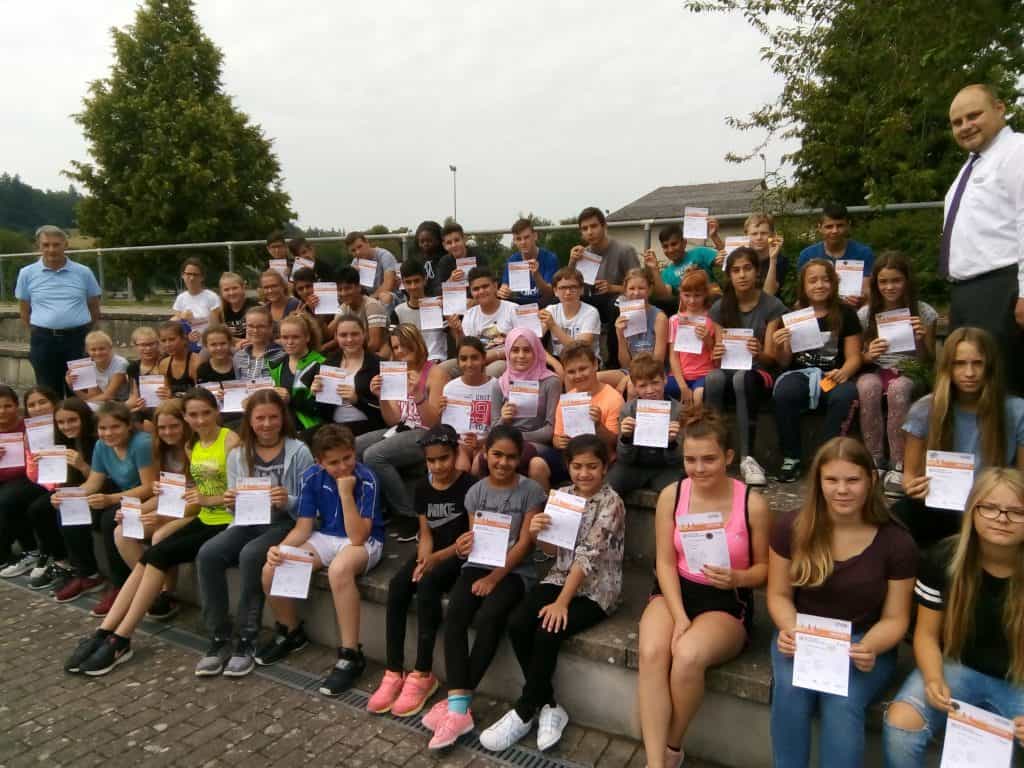 Eine große Gruppe von Kindern und zwei Erwachsenen posiert draußen auf Stufen, hält Zertifikate in den Händen und lächelt in die Kamera. Im Hintergrund sind Bäume und ein Zaun zu sehen.