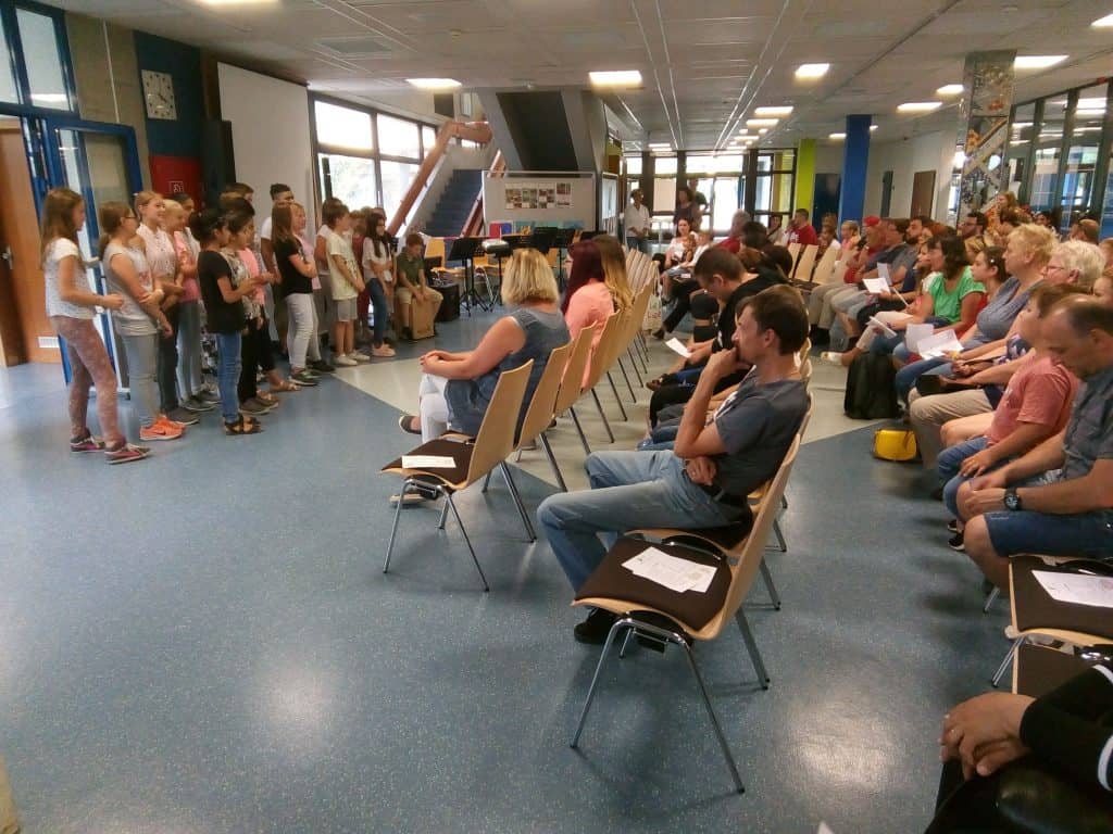 Eine Gruppe von Kindern steht vor sitzenden Erwachsenen in einem hellen Raum, möglicherweise einer Schule. Die Erwachsenen scheinen aufmerksam zuzuhören und halten Papiere in den Händen, während die Kinder scheinbar einen Vortrag halten oder eine Aufführung machen.