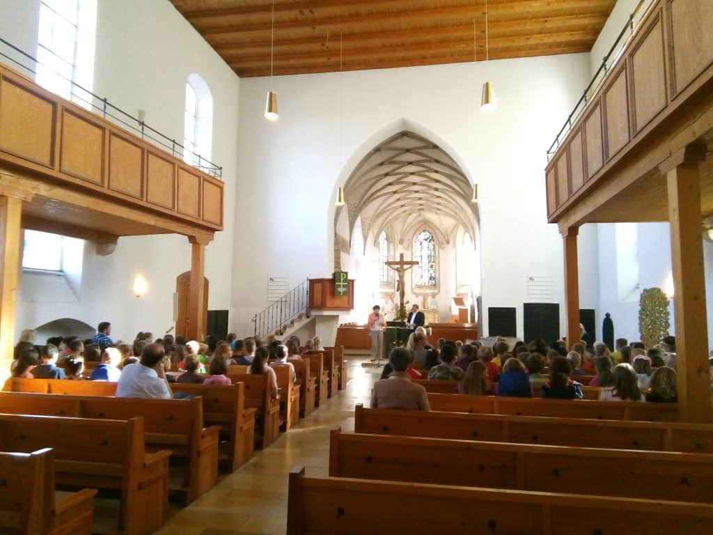 Eine Gemeinde sitzt in Holzbänken in einer hellen Kirche mit hohen Decken, gewölbter Architektur und einem großen Kreuz an der Vorderseite. Sonnenlicht strömt durch hohe Fenster und erhellt den Innenraum.